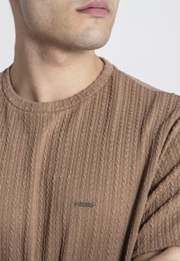 Bruine geribde gebreide trui met een ronde hals en korte mouwen. Bevat een klein merketiket aan de voorkant, dat een textuurdesign van stof toont.