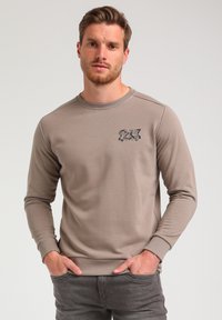 Light brown long-sleeve sweatshirt featuring a crew neck, ribbed cuffs, and a graphic logo on the left chest. Soft texture.