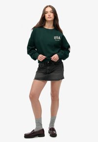 Sudadera verde con "USA" impreso en blanco, combinada con una falda de mezclilla oscura. El modelo lleva calcetines grises y mocasines burdeos.
