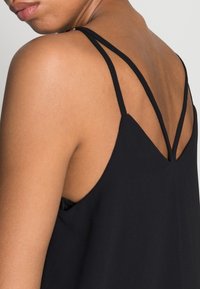 Canotta nera con spalline sottili, caratterizzata da un design con scollatura a V profonda e una texture liscia, che mette in risalto una silhouette minimalista e moderna.