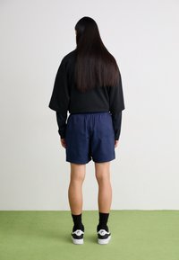 Black oversized sweatshirt, navy shorts, black socks, and white sneakers. Hair is long and straight. Standing on green flooring.