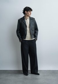 Jonge man met donker krullend haar staat met zijn handen in zijn zakken, draagt een zwart leren jack, beige polo, wijde zwarte broek en zwarte schoenen.