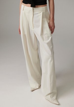 Femme portant un pantalon large crème et des chaussures blanches pointues, debout devant un fond gris uni.