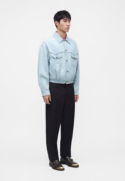 Homme portant une veste en jean bleu clair, un pantalon noir et des mocassins noirs avec bouts dorés, debout devant un fond blanc uni.