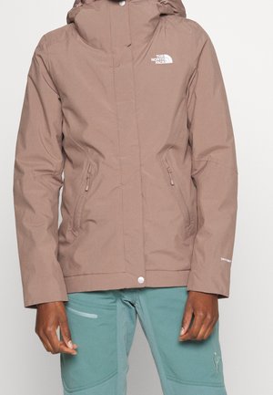 Vorderansicht einer Person, die eine braune North Face Jacke mit Reißverschlusstaschen und türkisfarbene Hosen mit einer Reißverschlusstasche am linken Oberschenkel trägt.