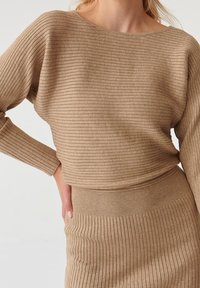 Kobieta w dopasowanym, prążkowanym, jasnobrązowym komplecie swetra i spódnicy z długimi rękawami oraz dopasowanym pasem, z ręką opartą na biodrze.