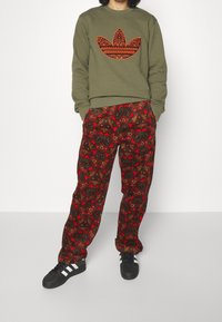 Olivgrön sweatshirt med orange blommigt logo, ihop med röda mönstrade byxor med blommotiv. Svarta sneakers med vita ränder.