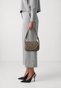 Sac à main marron à motifs avec une chaîne et une poignée en cuir, associé à une tenue à carreaux gris et blanc et des escarpins à talons retournés noirs.