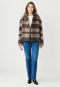 Veste marron à carreaux croisée avec un large col, détails de boutons et tissu texturé ; associée à un jean bleu et des chaussures noires.
