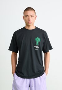 Mann som har på seg svart T-skjorte med grønn kaktusgrafikk og tekst, sammen med lyse lilla shorts, står mot en ensfarget hvit bakgrunn.
