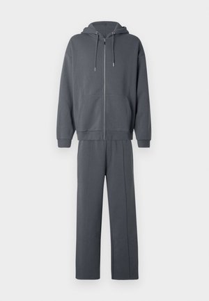 Tumši pelēks rāvējslēdzēja džemperis ar kabatām priekšpusē un atbilstošas taisnas bikses, attēlotas pret vienkāršu baltu fonu.