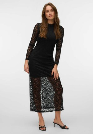 Femme debout portant une longue robe en dentelle noire avec des manches et un ourlet floraux transparents, associée à des sandales à talons ouvertes noires sur un fond uni.