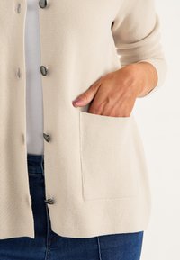 Beige stickad cardigan med en texturerad tyg, åtta runda knappar och en sidficka. Bärs över en vit topp och blå jeans.