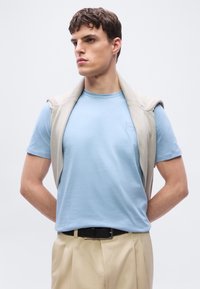 Νεαρός άνδρας που φορά ανοιχτό γαλάζιο μπλουζάκι, μπεζ πουλόβερ περασμένο στους ώμους, μπεζ παντελόνι και μαύρη ζώνη, στραμμένος αριστερά με τα χέρια πίσω από την πλάτη.