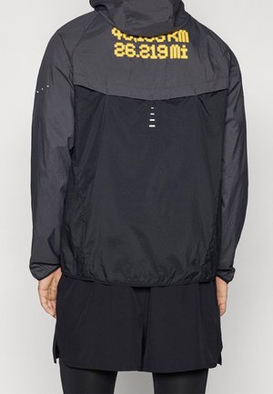 Veste noire à capuche avec texte jaune affichant "42.195 KM" et "26.219 Mi" sur le haut du dos, portée par une personne de dos.