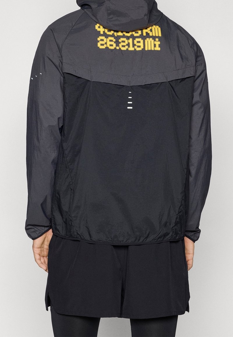 Veste noire à capuche avec texte jaune affichant "42.195 KM" et "26.219 Mi" sur le haut du dos, portée par une personne de dos.