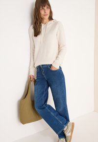 Hoodie crème texturé avec cordon de serrage ; jeans bleus taille haute ; sac fourre-tout vert olive ; baskets grises claires avec accents.