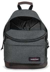 Mochila de tela gris con forro negro, fondo de cuero marrón y parche de logo. Cuenta con un asa superior y compartimentos con cremallera.