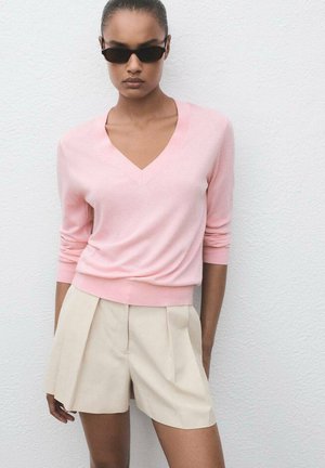 Lichtroze v-hals trui met lange mouwen, gecombineerd met beige geplooide shorts. Soepele textuur, ontspannen pasvorm, met een zoom die op de taille zit.