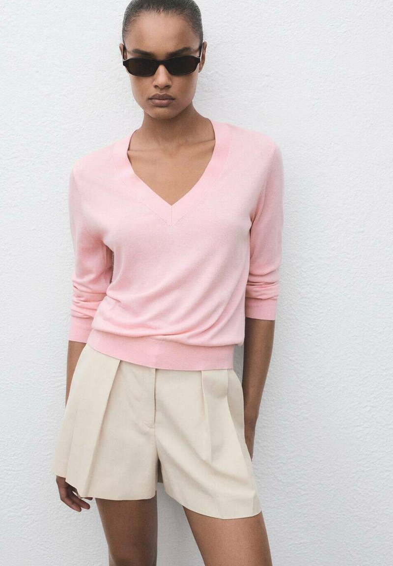 Lichtroze v-hals trui met lange mouwen, gecombineerd met beige geplooide shorts. Soepele textuur, ontspannen pasvorm, met een zoom die op de taille zit.