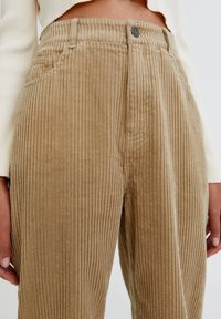 Pantalon en velours côtelé de couleur beige, présentant une texture côtelée verticale, des poches avant, une fermeture à bouton et une taille standard.