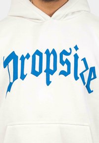 Hvid hættetrøje med blå gotisk skrift, der står "dropsize" på tværs af brystet, båret af en person.