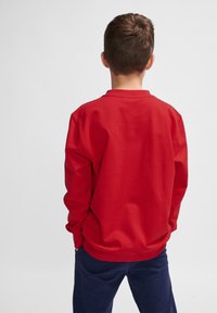 Felpa rossa con maniche lunghe e scollo rotondo. Presenta una texture liscia e polsini a coste. Indossata con pantaloni blu navy.