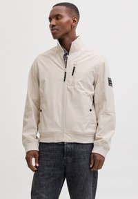 Veste zippée beige avec un col montant, deux poches poitrine zippées et des poches latérales à pression ; munie de poignets et d'un ourlet côtelés.