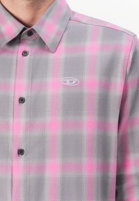 Camisa de botones en cuadros grises y rosas, con un logo gris claro, botones negros y un cuello estructurado; fabricada con una tela suave.