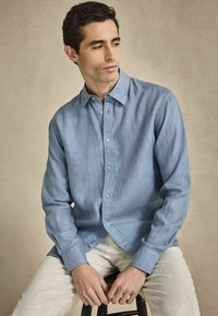Homme assis sur un tabouret portant une chemise bleu clair à boutons et un pantalon blanc, regardant vers sa gauche sur un fond uni.