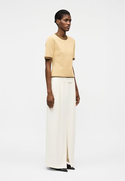 Kvinne som har på seg en kortermet beige rutete topp med brun kant og høytlivede vide hvite bukser med en frontsplitt.