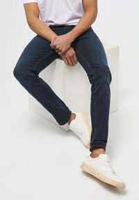 Lieknos tamsiai mėlynos džinsinės kelnės su parūkytomis apačiomis, derinamos su baltais odiniais sportbačiais, turinčiais kreminę padą, ant neutralaus fono.