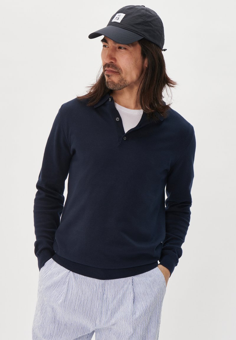 Dunkelblaues Strickpullover mit Kragen und Dreiknopf-Leiste. Lange Ärmel und gerippte Bündchen sowie Saum. Kombiniert mit hell gestreiften Hosen.