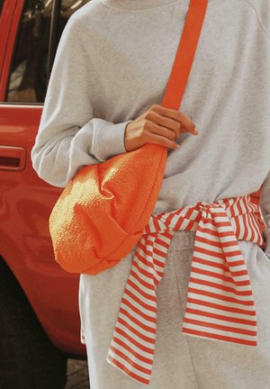 Oseba v svetlo sivi obleki z rdeče-belo črtasto srajco, zavezano okoli pasu, drži teksturirano oranžno torbico čez ramo blizu rdečega vozila.