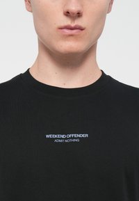 Zwarte katoenen T-shirt met een ronde halslijn, korte mouwen en witte geborduurde tekst met "WEEKEND OFFENDER ADMIT NOTHING" op de voorkant.