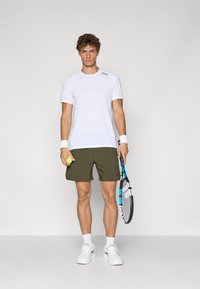 Camisola atlética de manga curta branca, calções verdes-oliva, segurando uma raquete de ténis e uma bola, usada com meias e sapatilhas brancas.