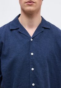 Άνδρας που φοράει πουκάμισο από λινό ναυτικό μπλε με κουμπιά και λαιμόκοψη τύπου κατασκήνωσης, εμφανιζόμενος από τους ώμους μέχρι τον κάτω λαιμό σε απλό φόντο.