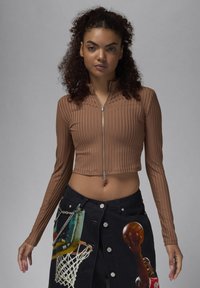 Brun ribbet crop-top med høy krage og frontglidelåsdetalj. Kombinert med et svart denimskjørt med grafiske utskrifter og sølvfargede detaljer.