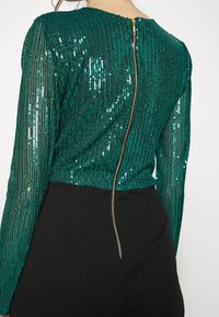 Top en sequins verts avec des rayures verticales, manches longues et une fermeture éclair dorée au dos. Associé à un pantalon taille haute noir.