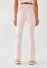 Jasnoróżowe legginsy rozkloszowane z wysokim stanem, gładką teksturą i modelującym krojem. Połączone z butami sportowymi w neutralnym kolorze.