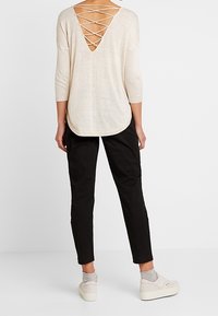 Beige stickad topp med djup V-back och korsningsdetalj, tillsammans med svarta avsmalnande byxor och ljusa sneakers.