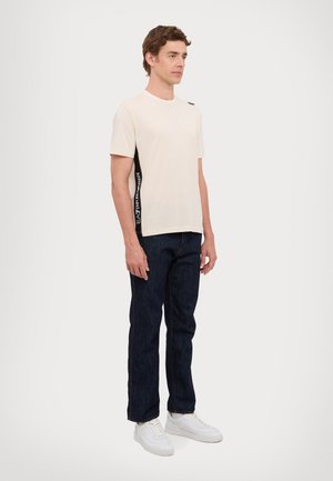 Beige cotton T-shirt with a crew neck, featuring a black side panel with white text. Paired with dark denim jeans and white sneakers.