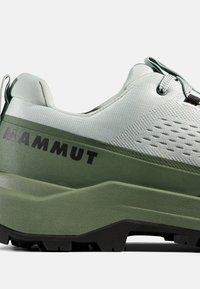 Jasnoszary i oliwkowozielony but trekkingowy z czarną podeszwą, oddychającą siateczkową cholewką oraz logo "MAMMUT" na boku.