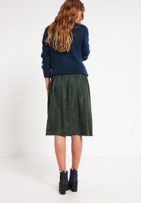 Suéter de punto azul marino, falda midi de ante verde, diseño acampanado, botas hasta el tobillo con tacones gruesos, textura suave y pliegues visibles en la falda.