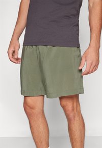 Mies, jolla on päällään oliivinvihreät shortsit ja tummanharmaa paita, seisoo kädet rentoina sivuilla tasaisen taustan edessä.