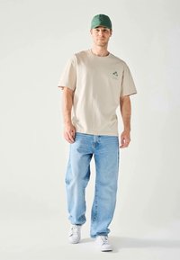 Homme portant un t-shirt beige avec un imprimé de palmier, un jean bleu clair, des baskets blanches et une casquette de baseball verte, se tenant devant un fond uni.