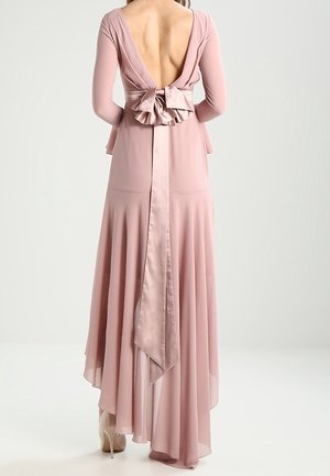 Femme portant une longue robe fluide rose poudré avec un dos ouvert et un grand détail nœud à la taille, associée à des talons hauts beiges.