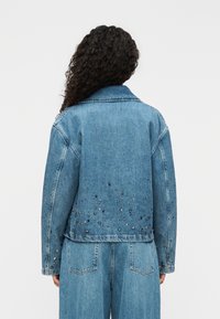 Τζιν μπουφάν μπλε χρώματος με κλασική γραμμή, διακοσμημένο με μεταλλικές πινέζες στο κάτω μέρος της πλάτης και με κομψό γιακά.