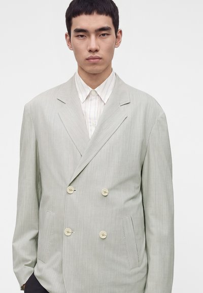 Jeune homme portant un blazer croisé gris clair sur une chemise blanche à col, debout devant un fond blanc uni.