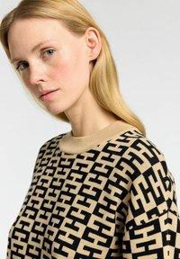 Beige neulepaita, jossa on musta geometrinen kuvio, pyöreä pääntie ja ribatulla yksityiskohdalla varustettu kaulus. Pehmeä neuletekstuuri on näkyvissä.